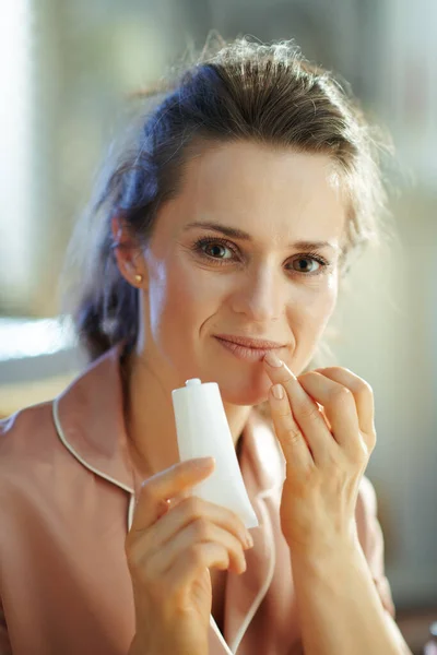 young woman in pajamas with white cosmetic tube applying lip contour cream in the modern house in sunny winter day.