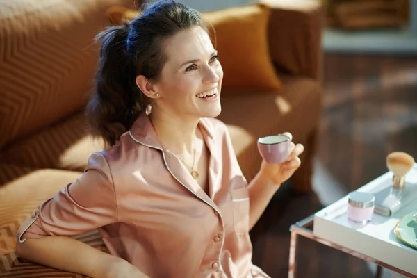 在阳光明媚的冬日里 穿着睡衣的快乐的现代女性在现代客厅的餐桌边喝咖啡和洗浴 — 图库照片