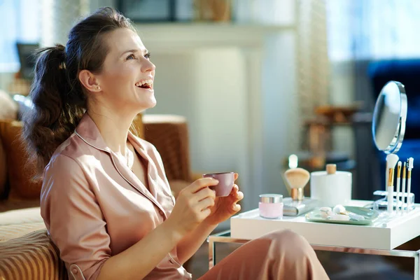 幸せな現代40歳の女性ですパジャマでコーヒーを飲みます近くのテーブルでバスアメニティ付き現代の家で晴れた冬の日 — ストック写真