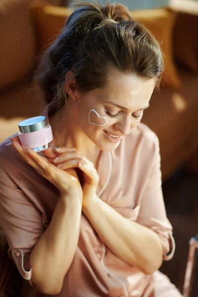 smiling elegant woman in pajamas with white facial cream heart on cheek holding pink cosmetic jar in the modern living room in sunny winter day.