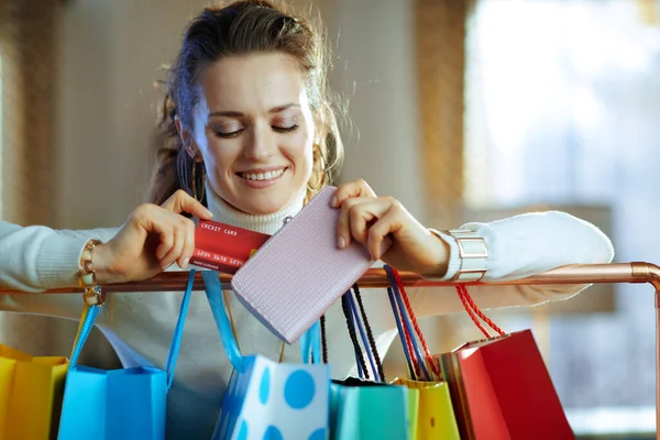 白いセーターとスカートの幸せな現代女性がカラフルなショッピングバッグの近くに銅製の服のレールにかかって財布からクレジットカードを取り出す — ストック写真