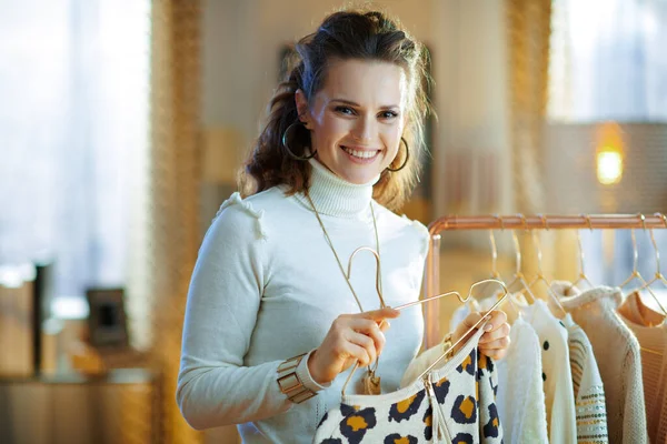 happy modern woman in white sweater and skirt at modern home in sunny winter day hanging sweater on a hanger near copper clothes rack.