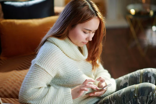 現代の10代の女の子とともに赤い髪で白いセーターで現代の家で晴れた冬の日の書き込みSms — ストック写真