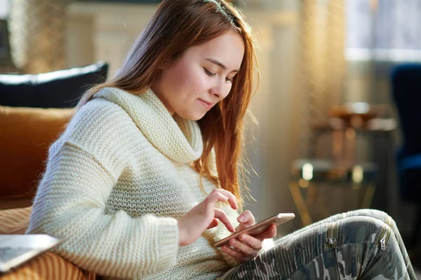 在阳光明媚的冬日里 身穿白色毛衣 头戴智能手机的快乐的现代女性推动着现代客厅社交媒体的发展 — 图库照片