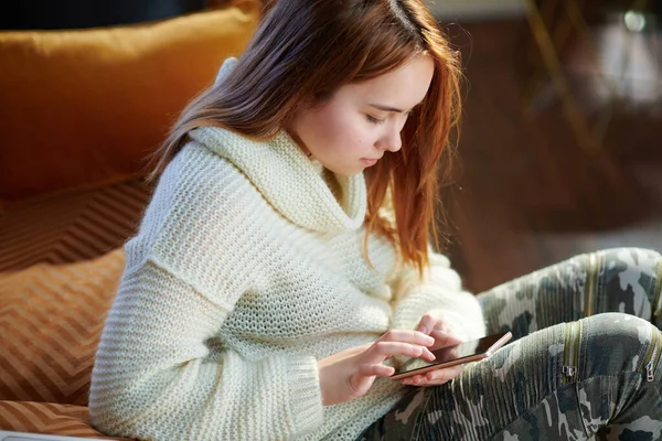 在阳光明媚的冬日里 穿着白色毛衣的现代少女在现代的房子里写下了Sms — 图库照片