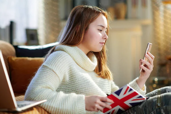 在阳光明媚的冬日里 身穿白色毛衣 头戴印有英国国旗的笔记本 手持智能手机在现代家中发短信的现代少女 — 图库照片