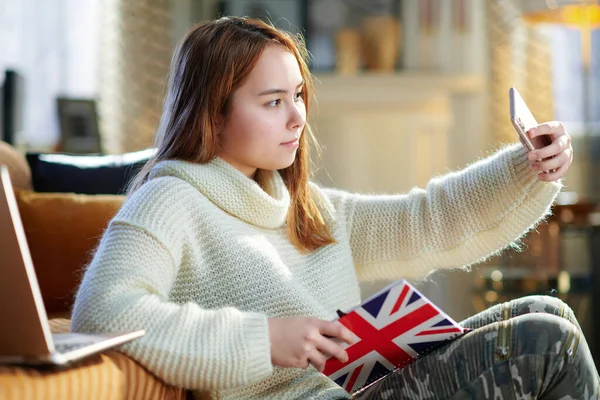 現代のティーン女の子で赤い髪で白いセーターでノートの色の英国の旗取りますセルフィーとともに携帯電話で現代の家で晴れた冬の日 — ストック写真