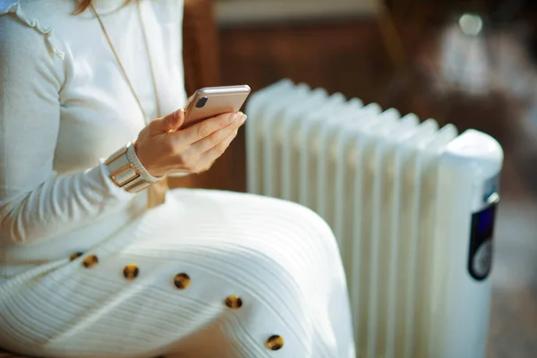 Modern woman in white sweater and skirt in the modern living room in sunny winter day sitting on couch near white electric oil radiator using smartphone app for temperature control.