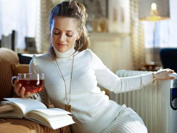 时尚的40岁家庭主妇 穿着白色毛衣和裙子 坐在沙发边和现代的白色电热炉旁 在阳光明媚的冬日里 在现代的房子里品着一杯热茶看书 — 图库照片