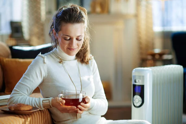 smiling modern woman in white sweater and skirt sitting near couch and white electric oil radiator with cup of hot tea and bun at modern home in sunny winter day.