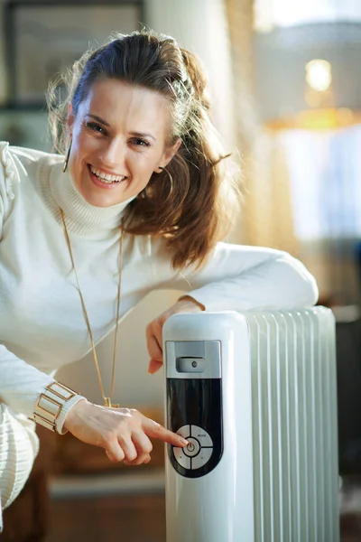 happy elegant middle age housewife in white sweater and skirt at modern home in sunny winter day turn on white electric oil radiator.