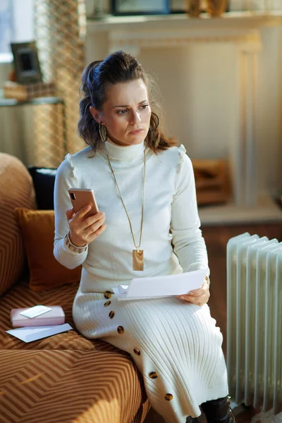 concerned young housewife in white sweater and skirt sitting on couch near white electric oil radiator with smartphone and utility bill in the modern living room in sunny winter day.