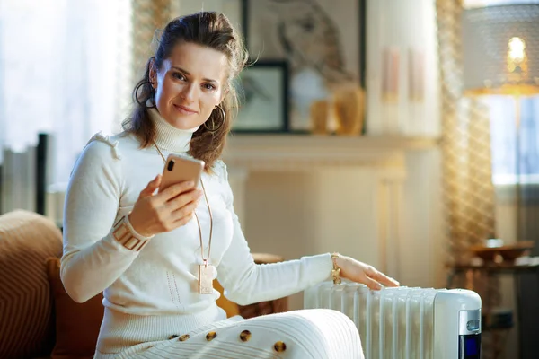 スタイリッシュな中年主婦白いセーターとスカートの現代の家で晴れた冬の日にソファの上に座っている温度制御のためのスマートフォンアプリを使用して白い電気オイルラジエーター — ストック写真