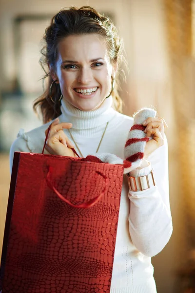 在阳光明媚的冬日里 身穿白色毛衣和裙子 头戴红色购物袋 穿着时髦的快乐女性形象在现代家中脱下采购的毛衣 — 图库照片