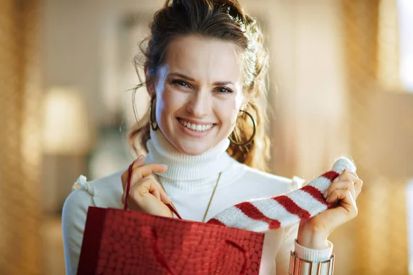 在阳光明媚的冬日里 穿着白色毛衣和裙子 头戴红色购物袋的优雅微笑的女人在现代客厅里拿出买来的毛衣的画像 — 图库照片