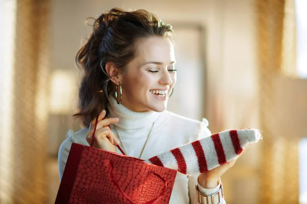 在阳光明媚的冬日里 身穿白色毛衣和裙子 头戴红色购物袋的快乐时尚女人在现代住宅里拿出她买的毛衣 — 图库照片