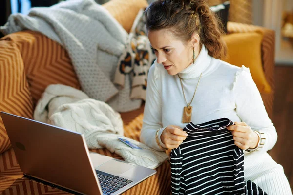young woman in white sweater and skirt with credit card sitting near couch among old sweaters while buying new sweater shop online on a laptop at modern home in sunny winter day.