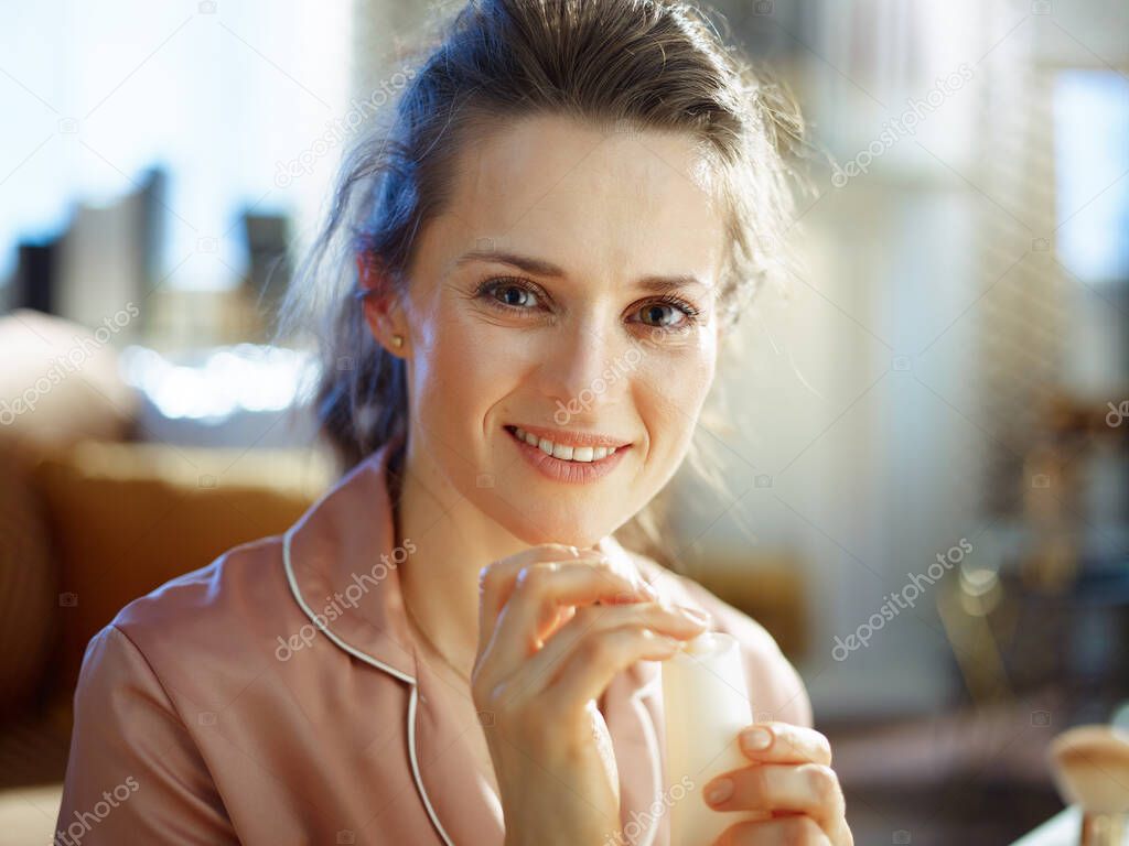 happy stylish middle age housewife in pajamas with white cosmetic tube at modern home in sunny winter day.