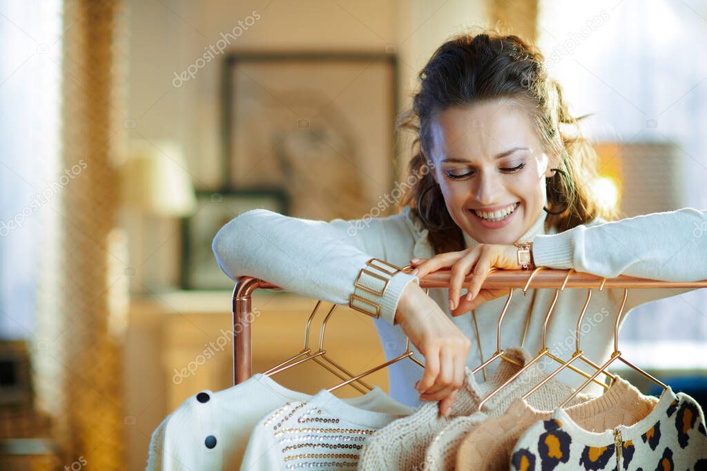 cheerful young woman in white sweater and skirt at modern home in sunny winter day choosing sweaters hanging on copper clothes rack.