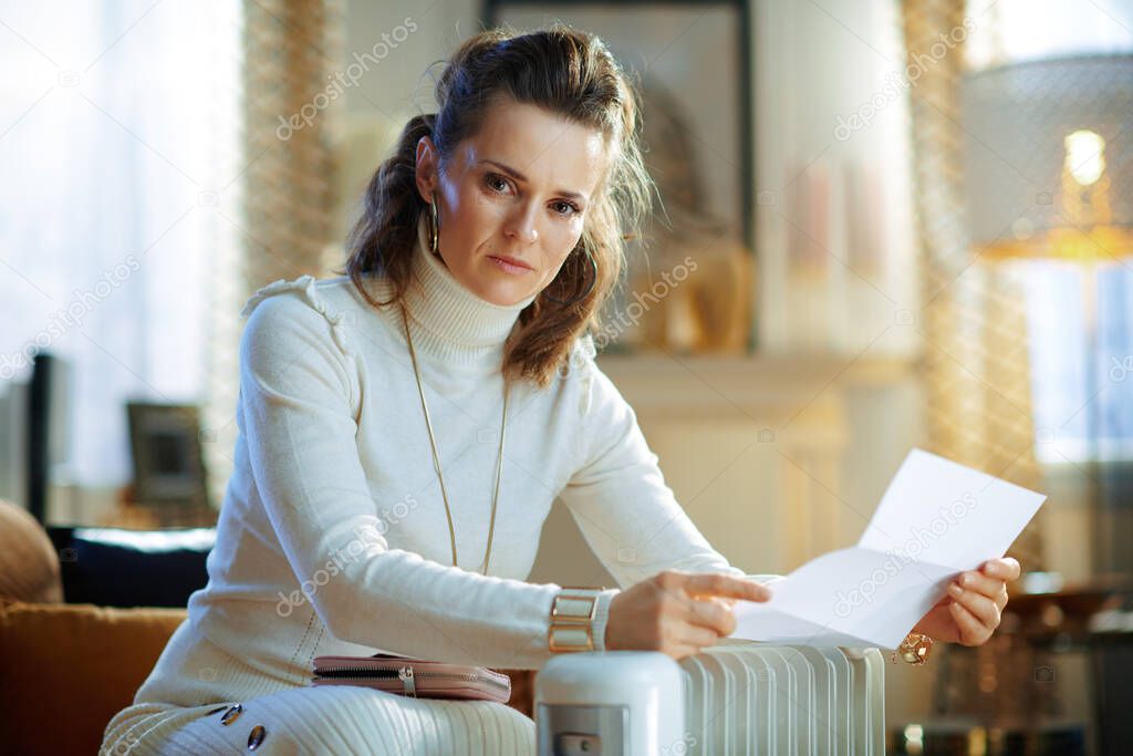 elegant female in white sweater and skirt sitting on couch with utility bill near white electric oil radiator at modern home in sunny winter day.