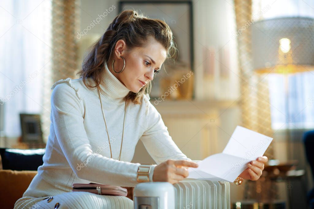 unhappy trendy housewife in white sweater and skirt sitting on couch with utility bill near white electric oil radiator in the modern living room in sunny winter day.