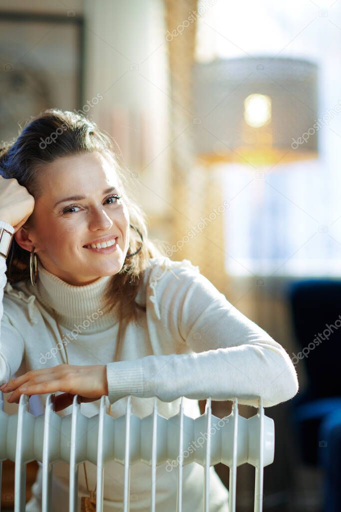 Portrait of smiling elegant middle age housewife in white sweater and skirt in the modern living room in sunny winter day near warm white electric oil radiator.