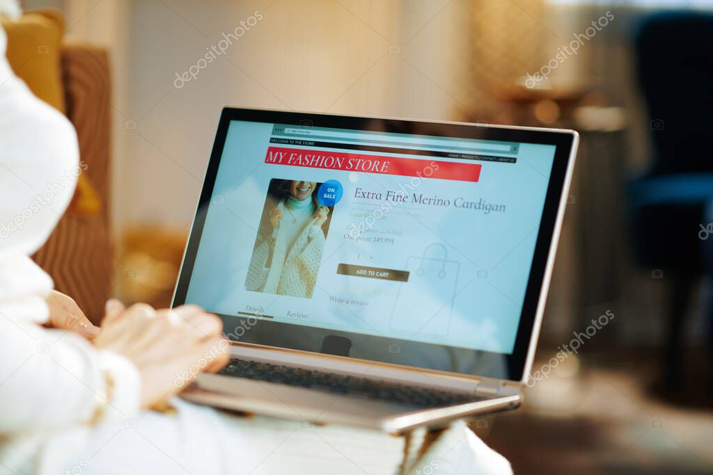 Closeup on modern woman in white sweater and skirt in the modern house in sunny winter day buying sweater while shop online on a laptop.