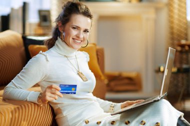 Beyaz kazaklı ve etekli gülümseyen modern kadın güneşli kış günlerinde modern evdeki e-ticaret sitesinden online alışveriş yapıyor..