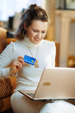 Beyaz kazaklı ve etekli gülümseyen modern kadın güneşli kış gününde modern oturma odasındaki e-ticaret sitesinde online alışveriş yapıyor..