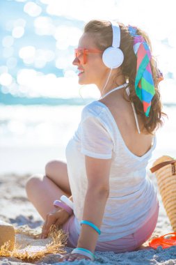 Beyaz tişörtlü ve pembe şortlu mutlu bir kadın deniz kenarında kulaklıkla müzik dinliyor..