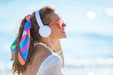 Beyaz tişörtlü, pembe şortlu, okyanus kıyısında kulaklıkla müzik dinleyen orta yaşlı bir kadının yan görüntüsü..