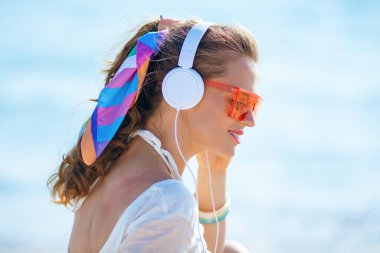 Beyaz tişörtlü, pembe şortlu, okyanus kıyısında kulaklıklı müzik dinleyen rahat modern bir kadın..