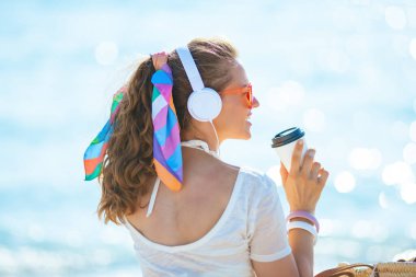 Gülümseyen 40 yaşındaki beyaz tişörtlü, pembe şortlu, kulaklıkla müzik dinleyen ve deniz kıyısında otururken kahve içen bir kadın..