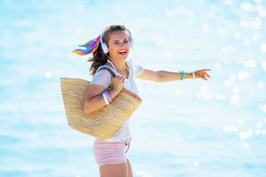 smiling young woman in white t-shirt and pink shorts with beach straw bag listening to the music with headphones and jumping on the beach. clipart
