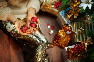 Süslü, orta yaşlı, altın pullu etekli, beyaz süveterli ev hanımı Noel ağacının altında elinde kırık bir Noel topu olan hediye kutularının yanında..