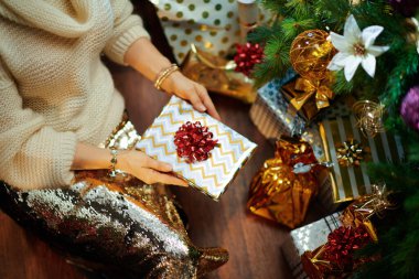 Süslü Noel ağacının altında altın pullu etekli ve beyaz süveterli zarif bir kadın, hediye kutularının yanında paketlenmiş kitap tutuyor..