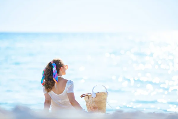 白いTシャツの現代の中世の女性の後ろから見たと海の海岸に座ってヘッドフォンやビーチストローバッグとピンクのショートパンツ — ストック写真