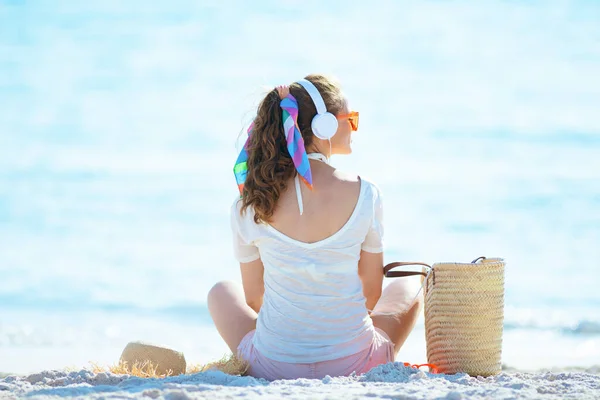 Seen from behind modern middle age woman in white t-shirt and pink shorts listening to music with headphones and sitting on the seashore.