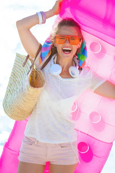 幸せな40歳の女性で白いTシャツとピンクのショートパンツでビーチストローバッグとともにインフレータブルマットレス ヘッドフォンとオレンジサングラスで海の海岸 — ストック写真