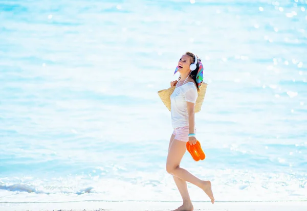 白いTシャツにピンクのショートパンツにオレンジのフリップフロップを持ったビーチストローのバッグを着て ヘッドフォンで音楽を聴きながら海岸を歩く流行の女性を笑顔 — ストック写真