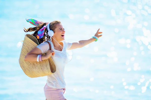 白いTシャツを着た陽気な若い女性とピンクのショートパンツとビーチストローバッグヘッドフォンで音楽を聞いて海の海岸に飛び込む — ストック写真