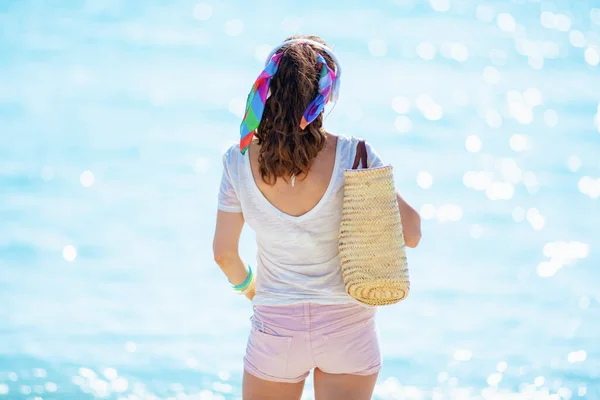 Seen from behind trendy woman in white t-shirt and pink shorts with beach straw bag listening to the music with headphones on the ocean shore.