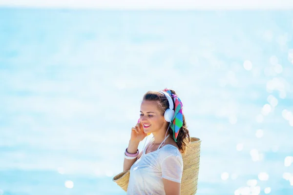 リラックスした健康的な女性で白いTシャツとピンクのショートパンツでビーチストローバッグで音楽を聴く海岸沿いのヘッドフォン — ストック写真