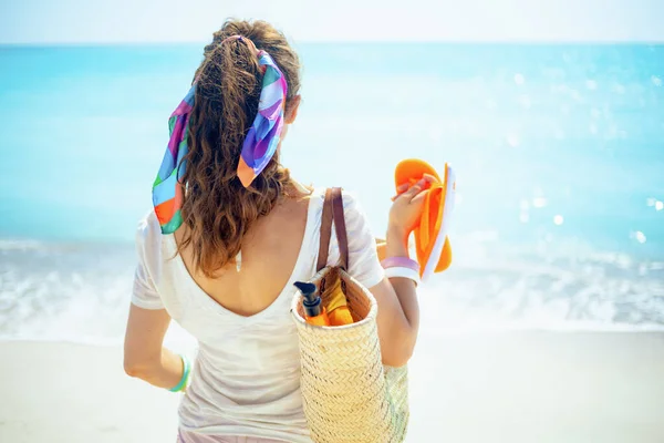 ビーチストローのバッグ オレンジのフリップフロップと海の海岸にスプフのボトルと白のTシャツの流行の40歳の女性の後ろから見た — ストック写真
