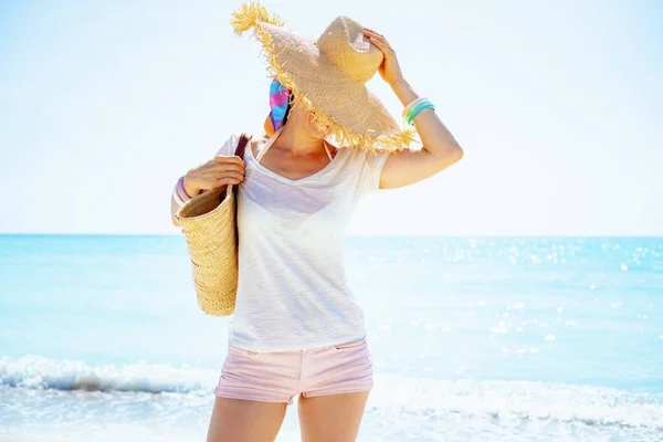 ビーチで大きな帽子をかぶっているビーチのわらの袋と白いTシャツの現代的な中世の女性 — ストック写真