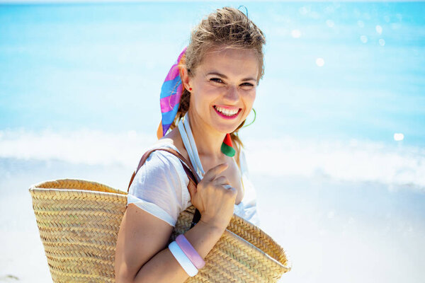 Portrait of happy trendy 40 year old woman in white t-shirt with beach straw bag on the ocean shore.