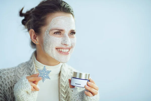 Happy woman in roll neck sweater and cardigan with white facial mask as part of winter skin care holding snowflake and cosmetic jar looking at copy space isolated on winter light blue background.