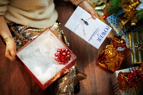 在装饰过的圣诞树下 穿着金色亮片裙和白色毛衣的时髦家庭主妇们在打开圣诞礼物包装的礼盒旁边穿上衣服 并获得了一本励志书籍 — 图库照片