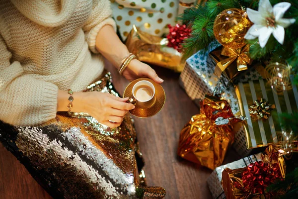 在装饰过的圣诞树下 穿着金色亮片裙和白色毛衣的中年妇女在礼品盒旁喝着一杯浓缩咖啡时穿的时髦衣服 — 图库照片