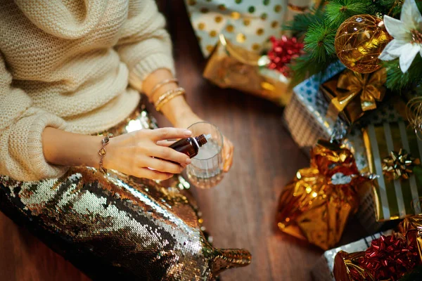 穿着金色紧身衣裙 头戴白色毛衣的优雅家庭主妇 在装饰圣诞树下的玻璃杯里 在礼物盒旁边滴着水 — 图库照片